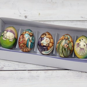 May include: A set of six decorative Easter eggs, each with a unique painted design. The eggs are displayed in a white box with individual compartments. The designs include religious scenes and a dove.