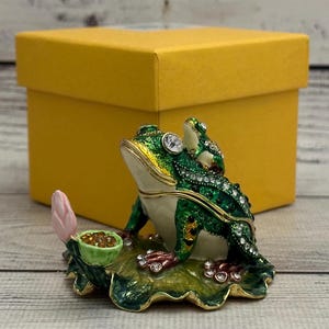 May include: A green frog figurine with a pink flower and a gold leaf, sitting on a green leaf. The frog is decorated with rhinestones and has a gold trim. The figurine is sitting on a wooden surface.