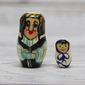 May include: Two hand-painted wooden nesting dolls. The larger doll is a woman with black hair, wearing a blue and white patterned dress. The smaller doll is a child with black hair, wearing a white dress with a blue collar.