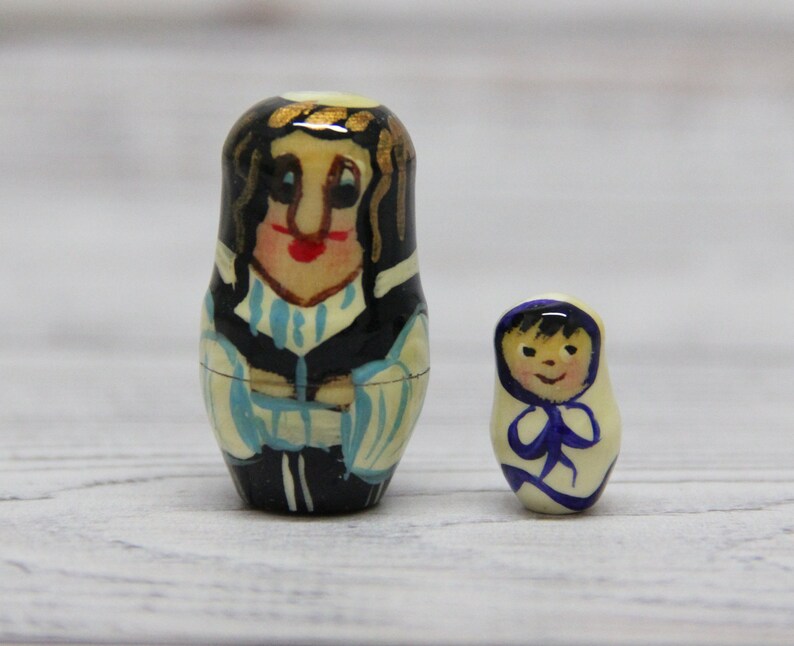 May include: Two hand-painted wooden nesting dolls. The larger doll is a woman with black hair, wearing a blue and white patterned dress. The smaller doll is a child with black hair, wearing a white dress with a blue collar.