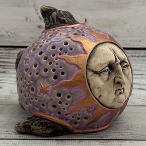 Könnte beinhalten: Eine kugelförmige Keramikskulptur mit einem modellierten menschlichen Gesicht mit ernstem Ausdruck. Die Oberfläche ist mit einem violetten Wolkenmuster, goldenen Sonnenstrahlen und kleinen sternförmigen Ausschnitten verziert. Dunkle, texturierte, felsartige Elemente befinden sich an der Ober- und Unterseite.