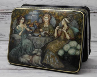 May include: A rectangular black box with a painted lid depicting three women at a table, a dog, and a garden scene. The women are dressed in long gowns, and the scene is rendered in soft, muted colors.