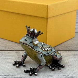 May include: A decorative frog figurine with a crown and jewel accents. The frog is painted in shades of green, gold, and purple. A yellow box is in the background. The frog is a unique decorative item.