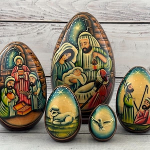 Peut inclure: Ensemble d'œufs de Pâques en bois peints à la main, représentant des scènes de la Nativité. Les œufs, de tailles variées, présentent des illustrations détaillées de personnages bibliques, dans un style rustique.