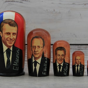 May include: A set of five Russian nesting dolls, or matryoshka dolls, featuring portraits of French presidents. The largest doll depicts Emmanuel Macron, followed by François Hollande, Nicolas Sarkozy, Jacques Chirac, and Émile Louis. Each doll is painted in a realistic style and has a unique design.
