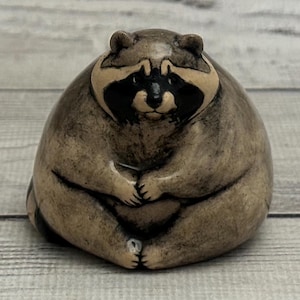Peut inclure: Une figurine en céramique d'un raton laveur assis avec ses pattes croisées devant lui. Le raton laveur est brun et gris avec des yeux noirs et un nez noir. La figurine est posée sur une surface en bois blanc.