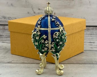 May include: A decorative egg-shaped trinket box featuring a deep blue enamel finish with gold-tone metal accents. The surface is adorned with green enameled lily of the valley leaves and clear crystal embellishments, topped with a small crown.
