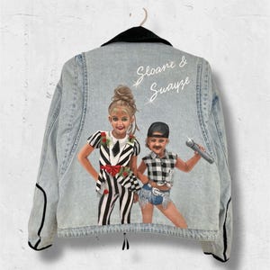 Peut inclure: Veste en jean bleu clair avec un col noir et des bordures noires. Le dos de la veste présente un portrait peint de deux enfants avec le texte "Sloane & Swayze" en écriture blanche. Un enfant porte une tenue rayée noire et blanche, et l'autre une chemise à carreaux.