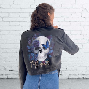 May include: Black leather jacket with a painted skull design on the back. The skull has one eye replaced with a blue butterfly and is surrounded by blue roses and leaves. The text "till death" is written in gold below the skull.