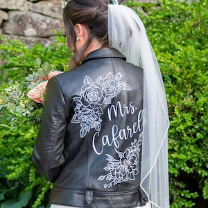 以下が含まれることがあります： 白い花の模様と「Mrs. Cafarelli」という文字が背中に書かれた黒い革のジャケット。白いベールをつけた人が着ています。