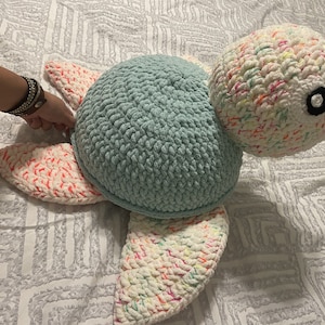 Puede incluir: Un gran peluche de tortuga hecho a mano con ganchillo. El cuerpo de la tortuga es de un azul claro suave, con una cabeza y aletas blancas con motas coloridas. Los bordes de las aletas son de color coral brillante. La tortuga tiene un ojo negro.