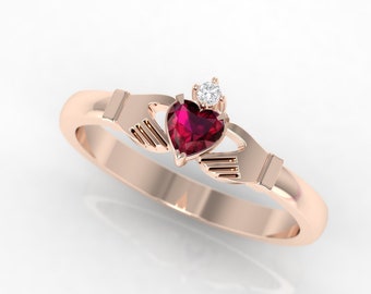 Puede incluir: Un anillo de oro rosa con un rubí en forma de corazón y un pequeño diamante de acento. El anillo presenta un diseño de nudo celta con dos manos que sujetan el corazón.