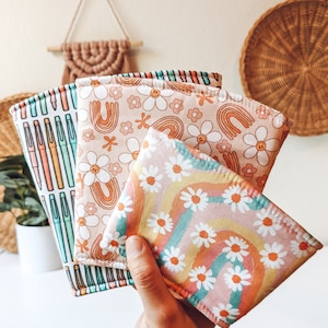 May include: Four colourful fabric cup sleeves with different patterns. The sleeves feature a rainbow, daisies, and a pattern of colourful pens.