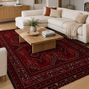 May include: A living room featuring a large, ornate, red area rug with a detailed pattern. The rug is centered beneath a light-colored sectional sofa. The room also includes a wooden coffee table, a wooden cabinet, and a neutral-toned painting.