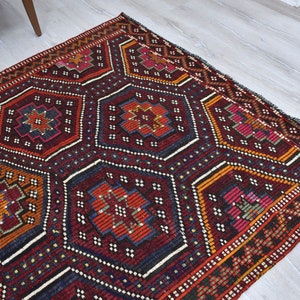 Pu&ograve; includere: Un tappeto turco tradizionale con un motivo geometrico in tonalit&agrave; di rosso, arancione, blu e bianco. Il tappeto ha uno sfondo scuro e un bordo dettagliato.
