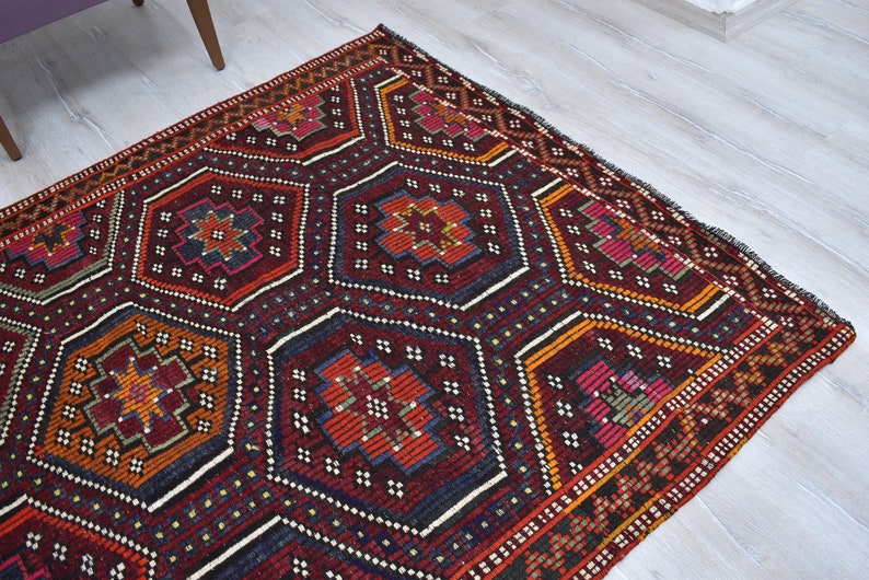 Pu&ograve; includere: Un tappeto turco tradizionale con un motivo geometrico in tonalit&agrave; di rosso, arancione, blu e bianco. Il tappeto ha uno sfondo scuro e un bordo dettagliato.