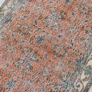 May include: A rectangular area rug featuring a distressed terracotta orange field with an intricate floral pattern in shades of navy blue, sage green, and cream. The rug has a wide decorative border with repeating geometric and botanical motifs.