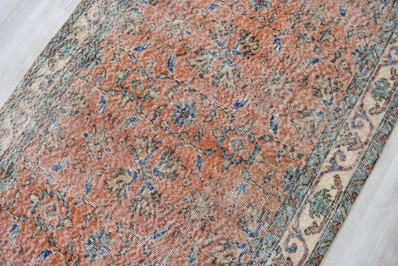 May include: A rectangular area rug featuring a distressed terracotta orange field with an intricate floral pattern in shades of navy blue, sage green, and cream. The rug has a wide decorative border with repeating geometric and botanical motifs.