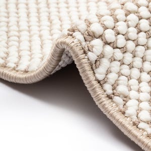 Può includere: Primo piano di un tappeto strutturato con un vello a ricciolo color crema e un bordo beige. Il design del tappeto presenta un motivo di piccole palline bianche in rilievo. Il bordo del tappeto è ripiegato, rivelando il lato inferiore.