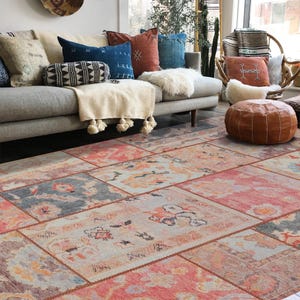 May include: A living room scene featuring a patchwork rug in shades of pink, orange, and gray. The rug is the focal point, complemented by a gray sofa adorned with various throw pillows in blue, rust, and cream. A leather ottoman and a woven chair complete the cozy setting.