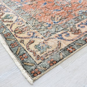 May include: A rectangular area rug featuring a central field of burnt orange with intricate floral and geometric patterns in blue, cream, and sage green. The border displays a repeating motif in dark green and cream with white fringe edges.