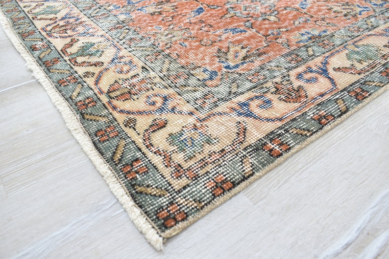 May include: A rectangular area rug featuring a central field of burnt orange with intricate floral and geometric patterns in blue, cream, and sage green. The border displays a repeating motif in dark green and cream with white fringe edges.
