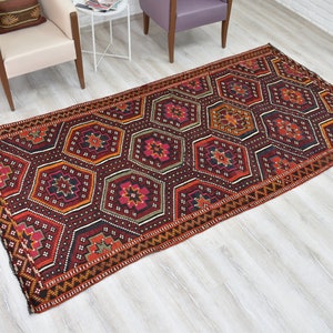 Pu&ograve; includere: Un tappeto rosso, arancione e nero con un design geometrico. Il tappeto ha un bordo con frange ed &egrave; steso su un pavimento in legno chiaro.