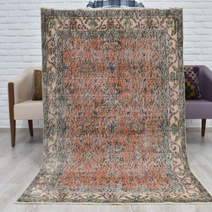 May include: A rectangular area rug featuring a central field of burnt orange with intricate floral and vine patterns in shades of blue, cream, and dark brown. The rug is framed by a wide, ornate border with repeating botanical motifs in similar colors.