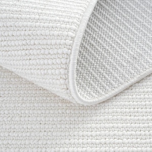 May include: Close-up of a white, textured bath mat with a looped pile. The edge is finished with a smooth, white border. The mat is folded over, revealing the underside with a dotted texture.