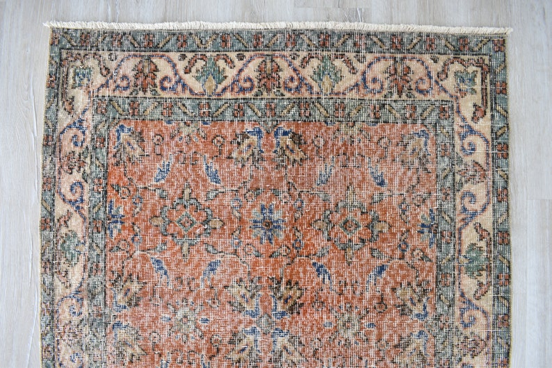 May include: A rectangular area rug featuring a central field of burnt orange with intricate floral and geometric patterns in shades of blue, green, and cream. The rug is framed by a wide decorative border and finished with a short white fringe.