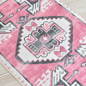 May include: Pink and white rug with a geometric pattern in black. The pattern features stylized birds and a central diamond shape.