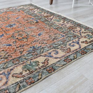 May include: A rectangular area rug featuring a central orange field with intricate floral patterns in blue, green, and cream. The rug has a wide decorative border with geometric and botanical motifs, finished with short white fringe on the edge.