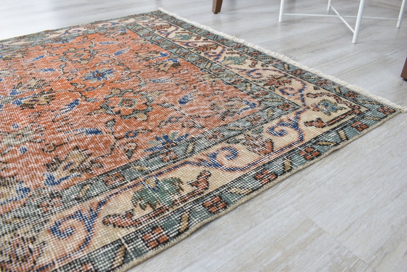 May include: A rectangular area rug featuring a central orange field with intricate floral patterns in blue, green, and cream. The rug has a wide decorative border with geometric and botanical motifs, finished with short white fringe on the edge.