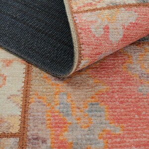 May include: A rectangular rug with a geometric pattern in shades of red, orange, gray, and beige. The rug's edge is visible, revealing a dark gray backing. The design features a patchwork style.