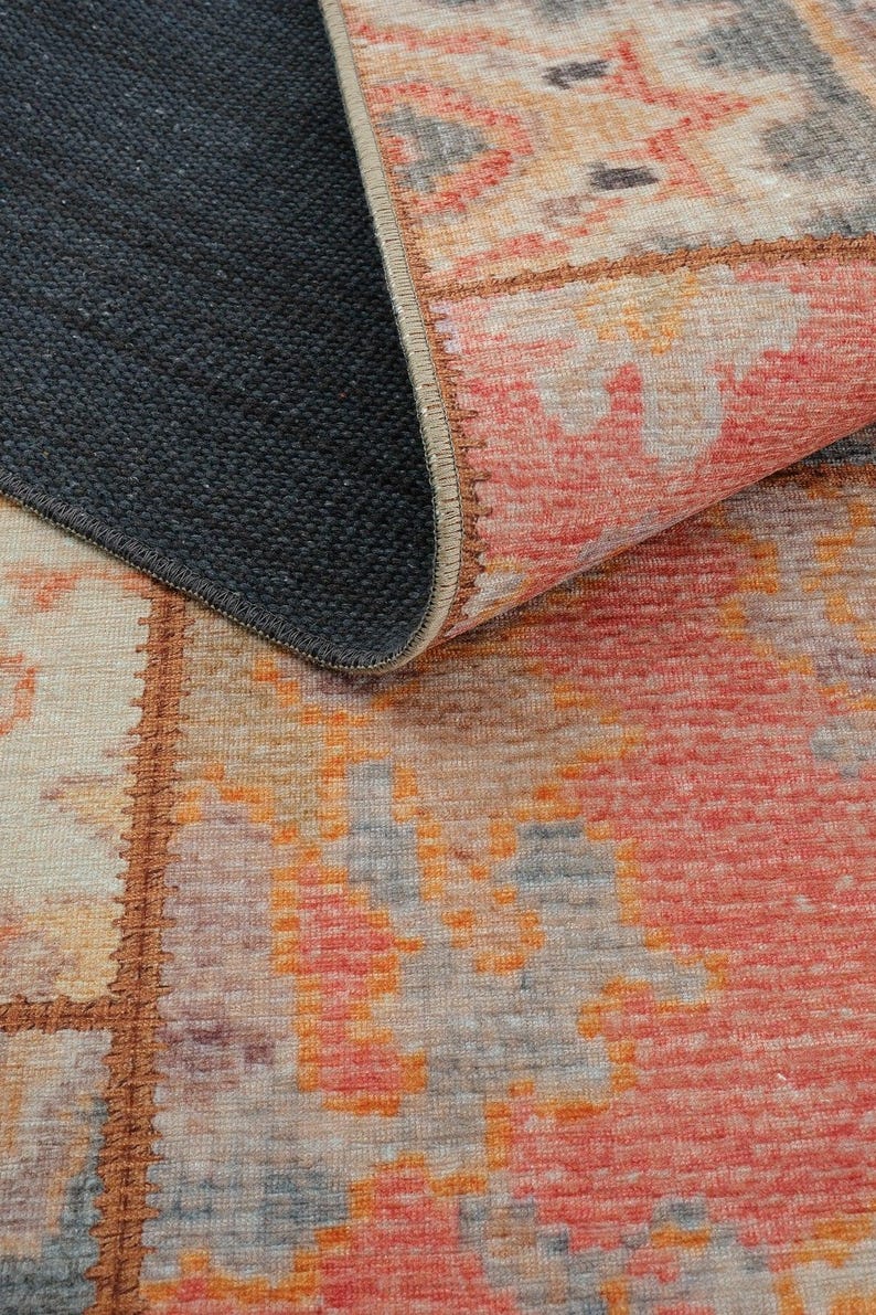 May include: A rectangular rug with a geometric pattern in shades of red, orange, gray, and beige. The rug's edge is visible, revealing a dark gray backing. The design features a patchwork style.