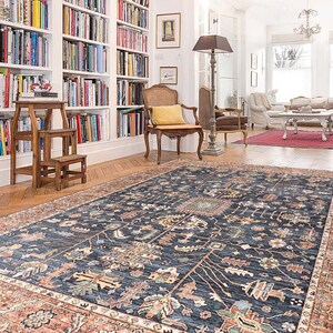 K&ouml;nnte beinhalten: Ein dunkelblauer Teppich mit traditionellem persischem Design. Der Teppich hat einen roten und cremefarbenen Rand und liegt auf einem Holzboden in einem Raum mit wei&szlig;en W&auml;nden und B&uuml;cherregalen.