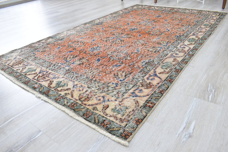 May include: A rectangular area rug featuring a central field of burnt orange with intricate floral and vine patterns in blue and cream. The wide border displays a repeating botanical motif in muted green, blue, and orange tones with fringe.