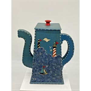 May include: A blue, hand-painted teapot with a nautical theme. The teapot features a red knob, a lighthouse design, and a sailboat. The waves are textured, and the teapot is decorated with white dots.
