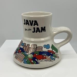 Puede incluir: Una taza de cerámica blanca con una base ancha y acampanada y un asa. El texto "JAVA for the JAM" está impreso en negro. Debajo del texto, un gráfico colorido representa un paisaje urbano con un atasco de coches rojos, azules y verdes.