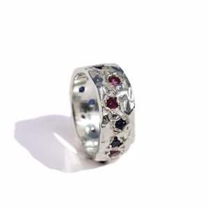 May include: A silver ring with a textured surface, adorned with multiple red and blue gemstones. The ring is set against a white background, showcasing its intricate design and craftsmanship. The gemstones are evenly spaced around the band.