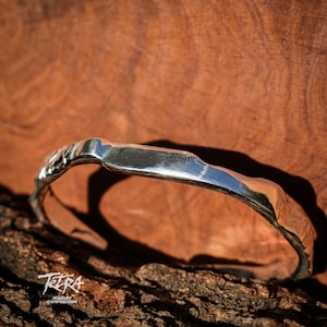 May include: A silver bracelet with a textured, organic shape. The bracelet is resting on a piece of wood.