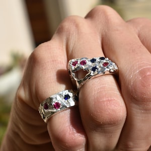 May include: Two silver rings with a textured design, each adorned with red and blue gemstones. The rings are held up close, showcasing the intricate details of the jewelry. The gemstones are set into the silver band.