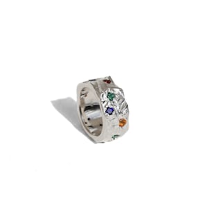 May include: A silver ring with a textured surface and colorful gemstones. The ring features a square shape and is adorned with red, green, blue, and orange stones. The ring is set against a white background.