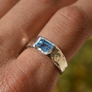 May include: A silver ring with a rectangular, light blue gemstone. The ring has a hammered texture and is worn on a finger. The gemstone is set in a bezel setting, and the ring appears to be handmade.