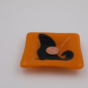 Op de afbeelding: Vierkant oranje glazen schaaltje met een zwart abstract ontwerp. Een koperen munt ligt in het schaaltje. Het schaaltje is een decoratief item.