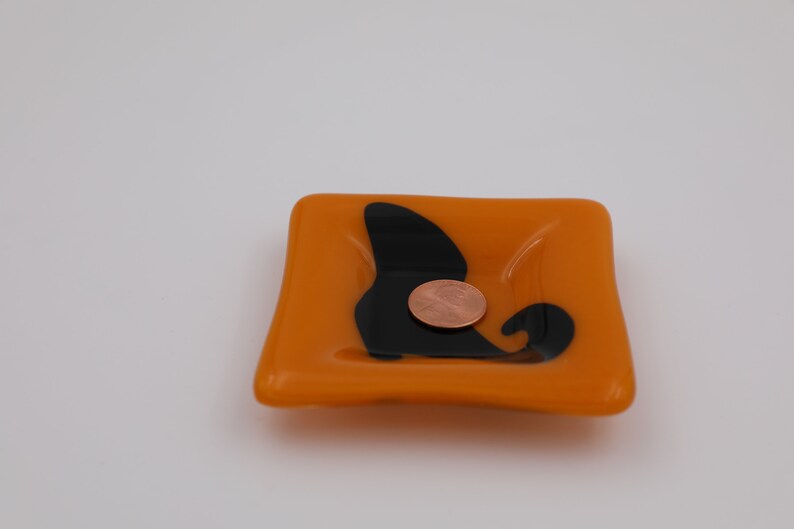 Op de afbeelding: Vierkant oranje glazen schaaltje met een zwart abstract ontwerp. Een koperen munt ligt in het schaaltje. Het schaaltje is een decoratief item.