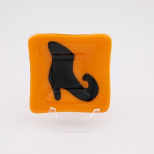 May include: A square, orange glass dish with a black witch's boot design. The dish is displayed on a clear plastic stand against a white background. The glass has a slightly wavy edge.