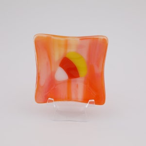 May include: A square, fused glass dish in shades of orange, yellow, and white. The design features a candy corn motif in the center. The dish is displayed on a clear plastic stand.