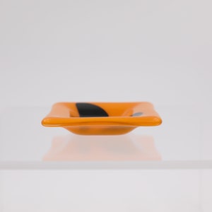 Op de afbeelding: Een kleine, vierkante oranje glazen schaal met een zwart abstract ontwerp. De schaal heeft licht gebogen zijkanten en staat op een wit oppervlak. Het ontwerp is een eenvoudig, modern accent.