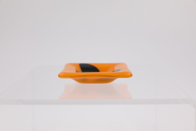 Op de afbeelding: Een kleine, vierkante oranje glazen schaal met een zwart abstract ontwerp. De schaal heeft licht gebogen zijkanten en staat op een wit oppervlak. Het ontwerp is een eenvoudig, modern accent.